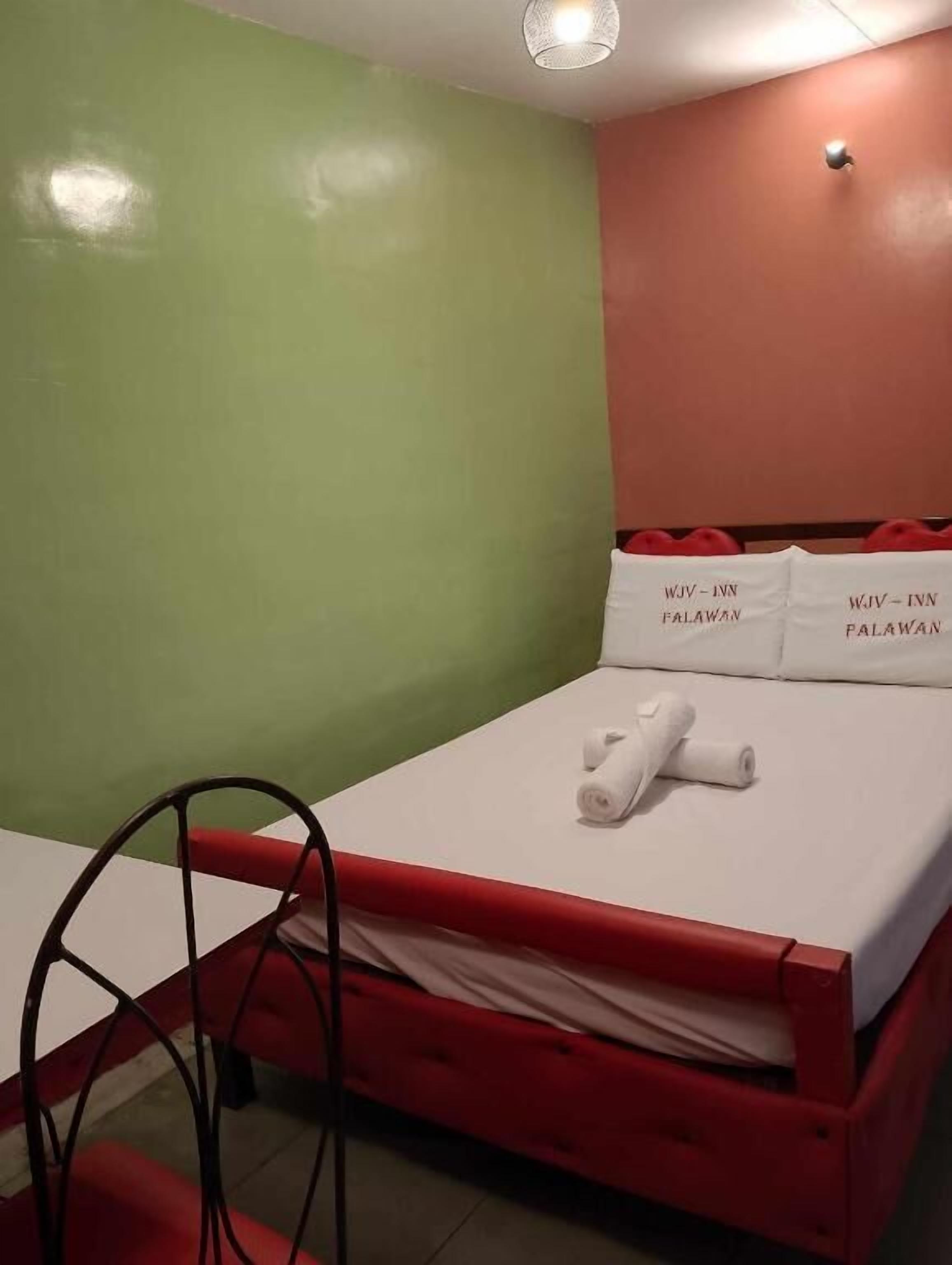Un lit simple avec un cadre rouge et du linge de lit blanc, un oreiller blanc avec l’inscription « WJIV - INN » et une serviette blanche roulée.