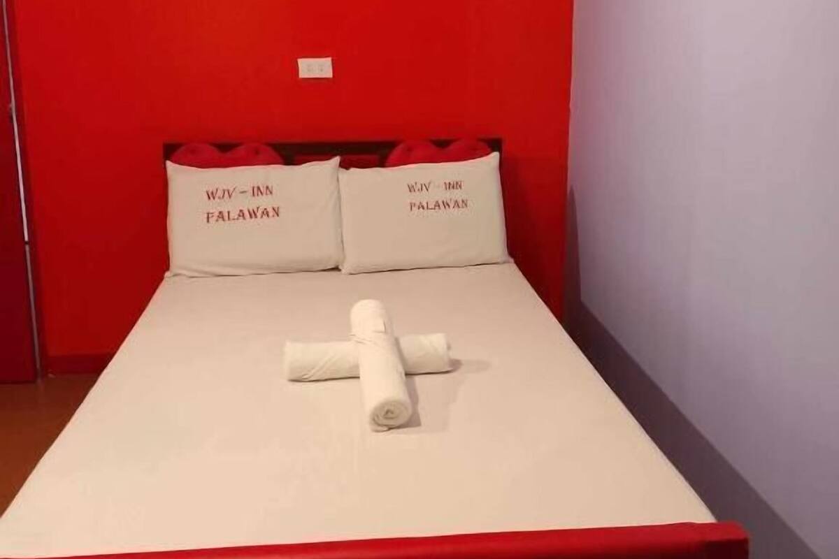 A small, red-walled room with a single bed, two pillows, and a rolled towel.