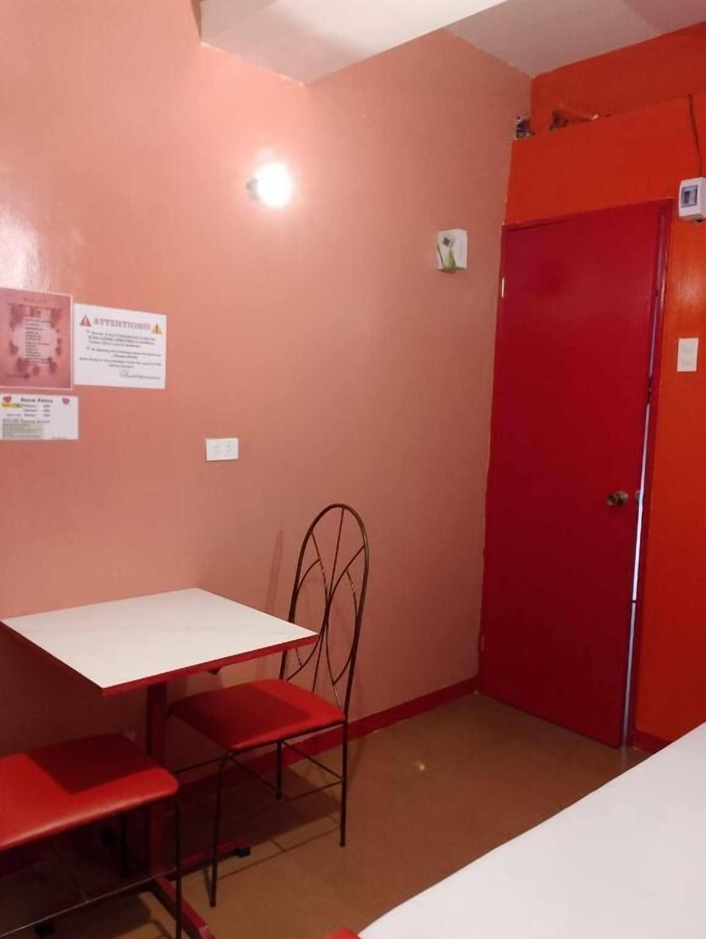 A small room with a red door, a white table, and a metal chair. There is a notice on the wall and a light fixture above the door.