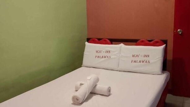Ett enkelrum med en röd sänggavel, gröna och orange väggar samt två kuddar med texten "WJIV - INN PALAWAN".