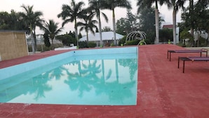 A swimming pool with a red deck, surrounded by palm trees and a covered area in the background. - Riyasat Panthera (Bali)