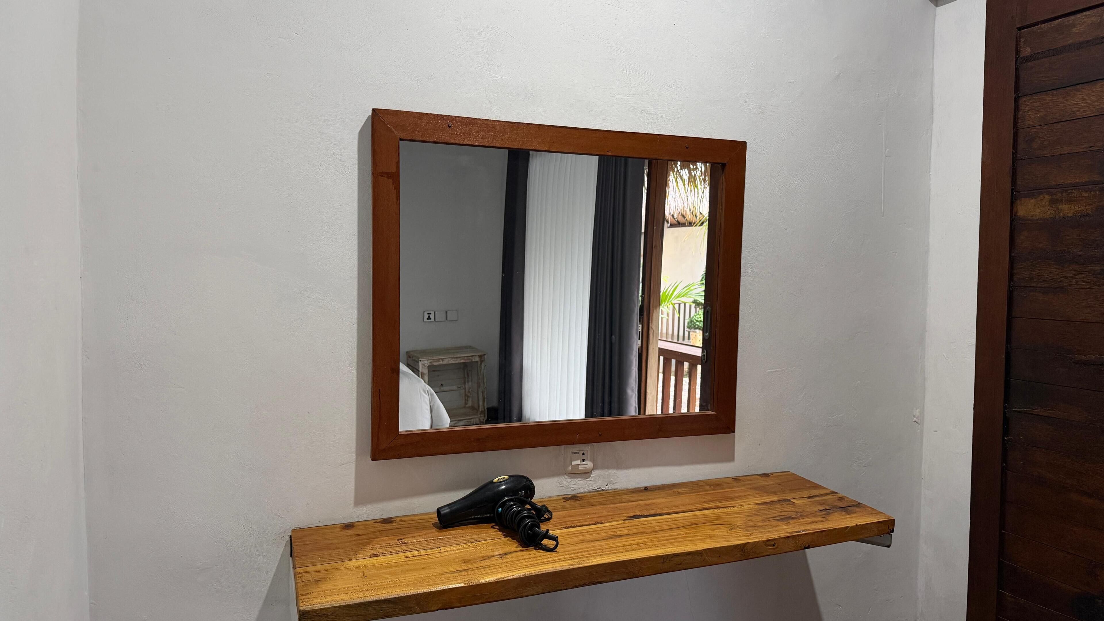 A mirror with a wooden frame mounted on a white wall, above a wooden shelf with a black object on it.