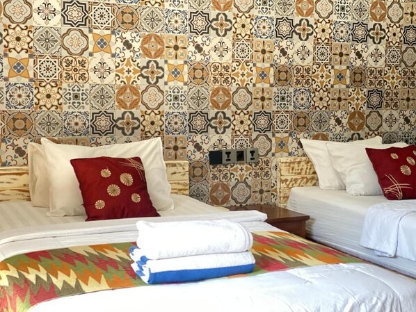 Habitación de hotel con dos camas, cada una con una almohada y una toalla blanca doblada con cuidado. Las paredes están decoradas con un diseño de azulejos estampados.