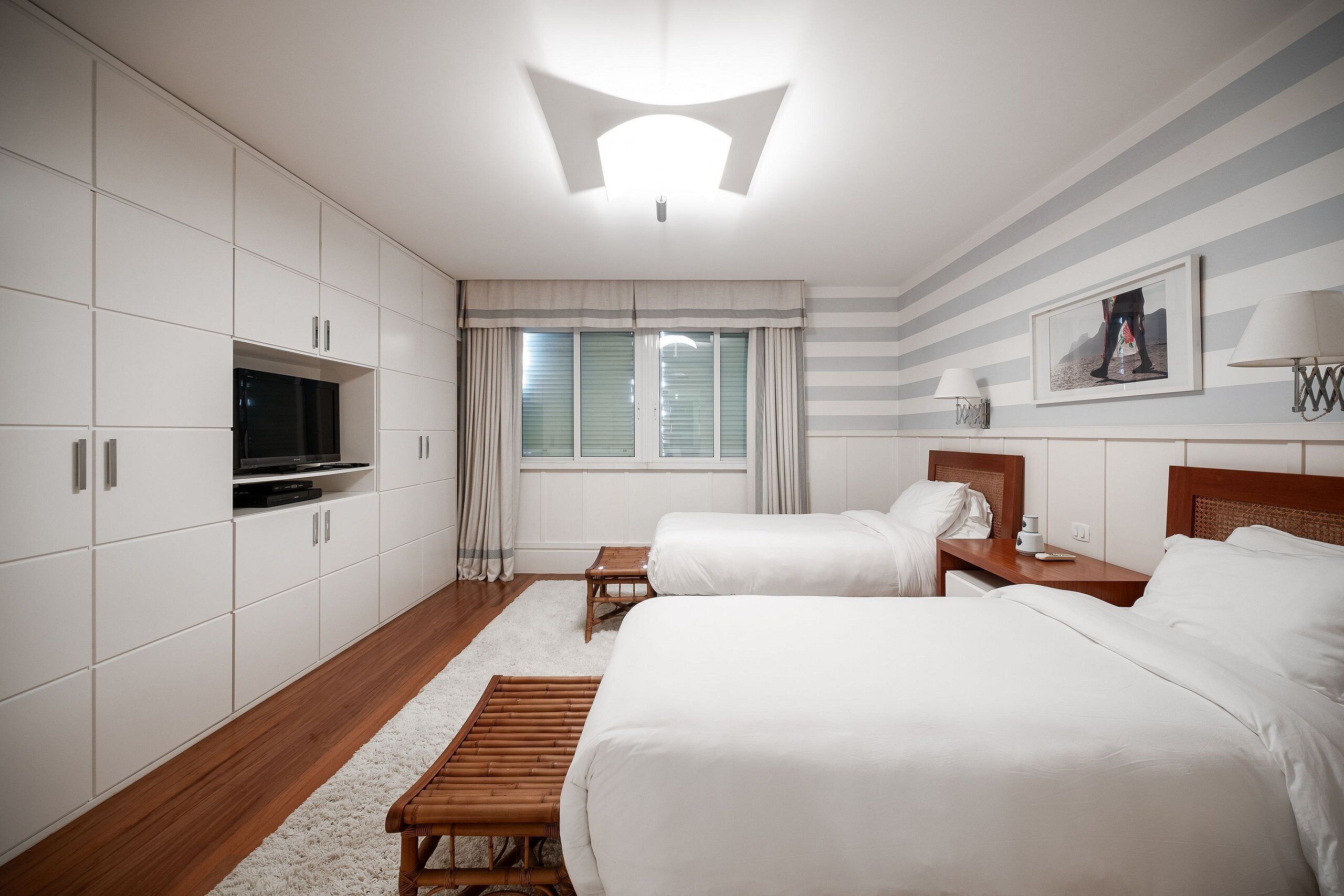 A hotel room with two beds, a TV, a wooden bench, a small table, and a window with curtains.