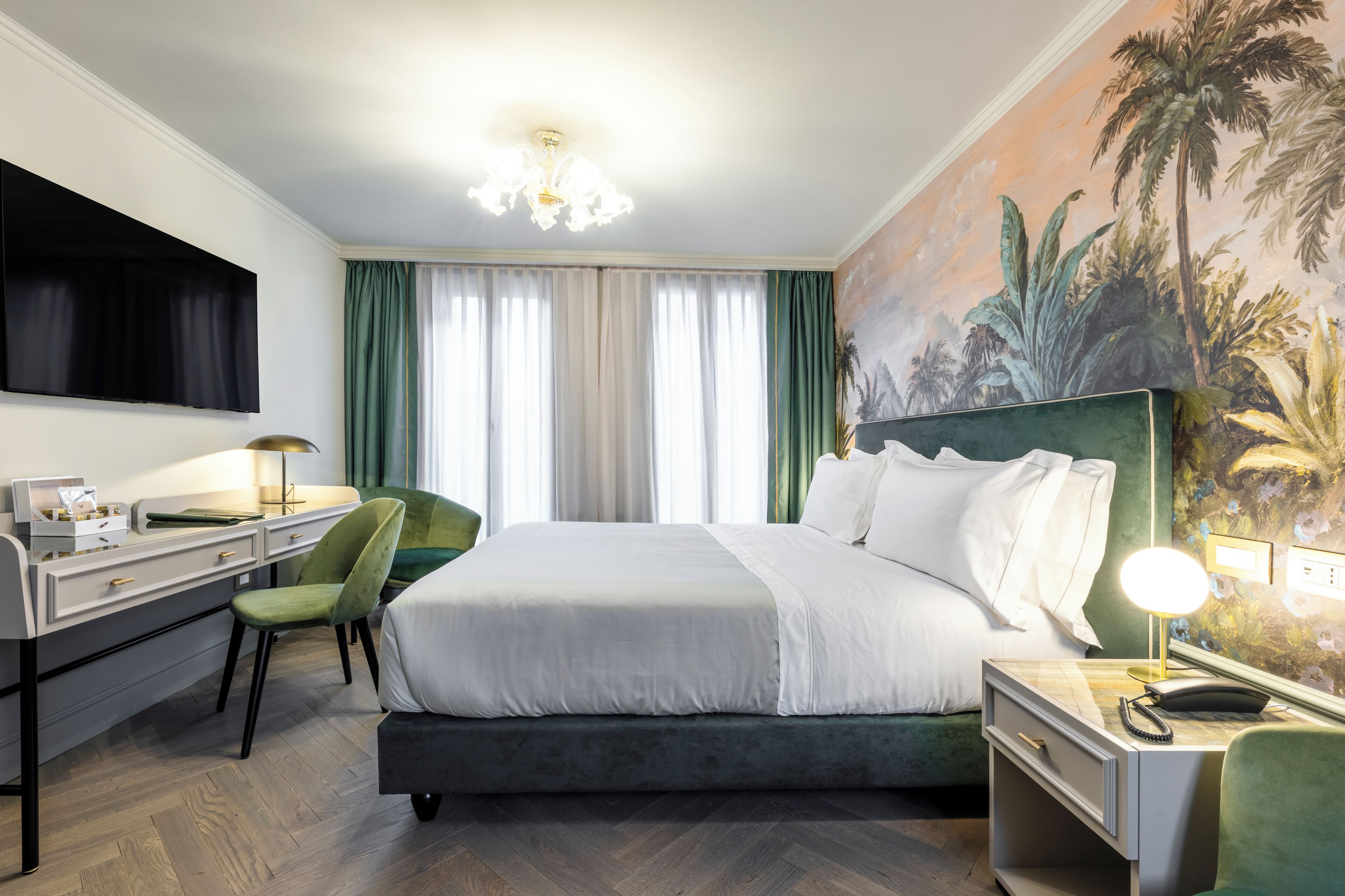 A hotel room with a large bed, a desk with a lamp, a chair, and a painting of tropical scenery on the wall.