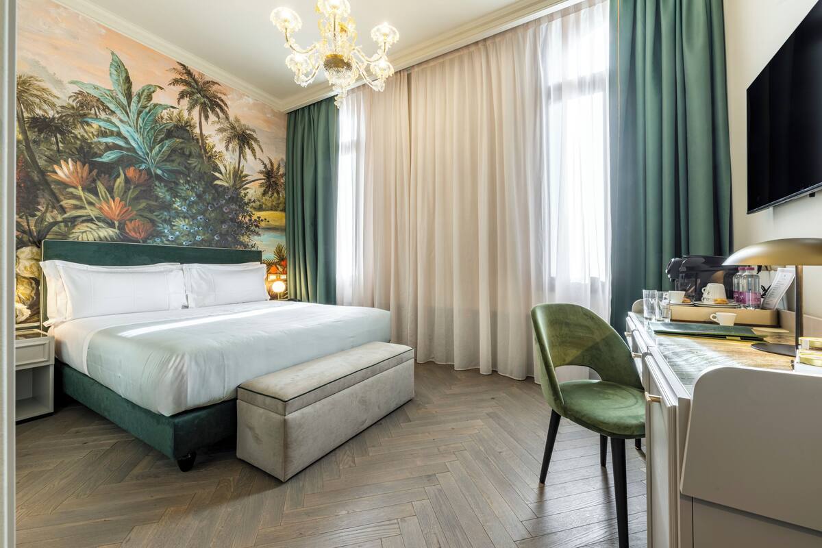 A hotel room with a bed, a desk with a chair, a television, and a large mural on the wall.