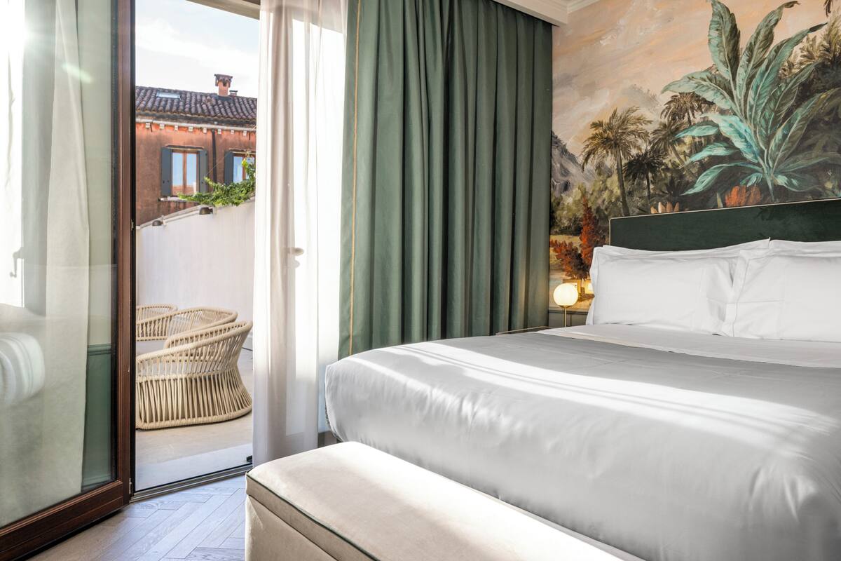 A hotel room with a bed, a view of a building through a glass door, and a mural of tropical foliage.
