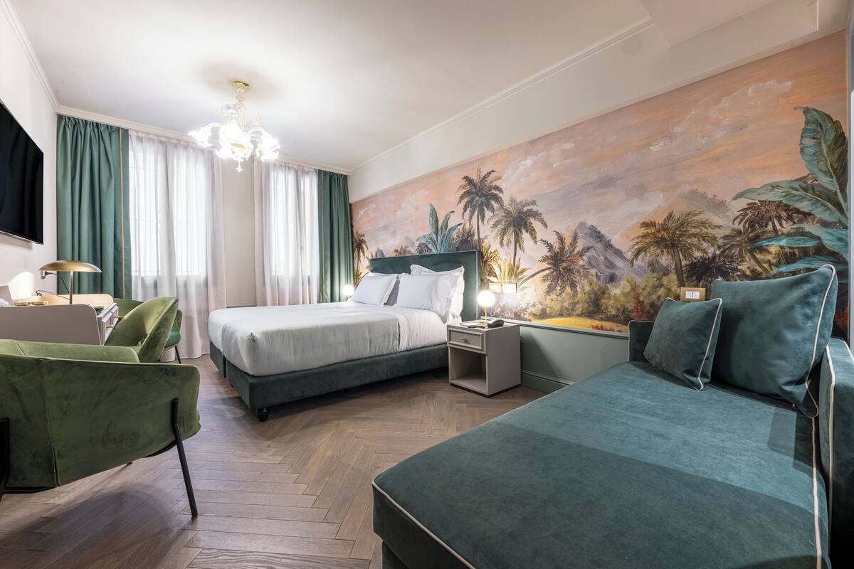 A hotel room with two beds, a green armchair, a desk with a lamp, and a mural of tropical scenery.
