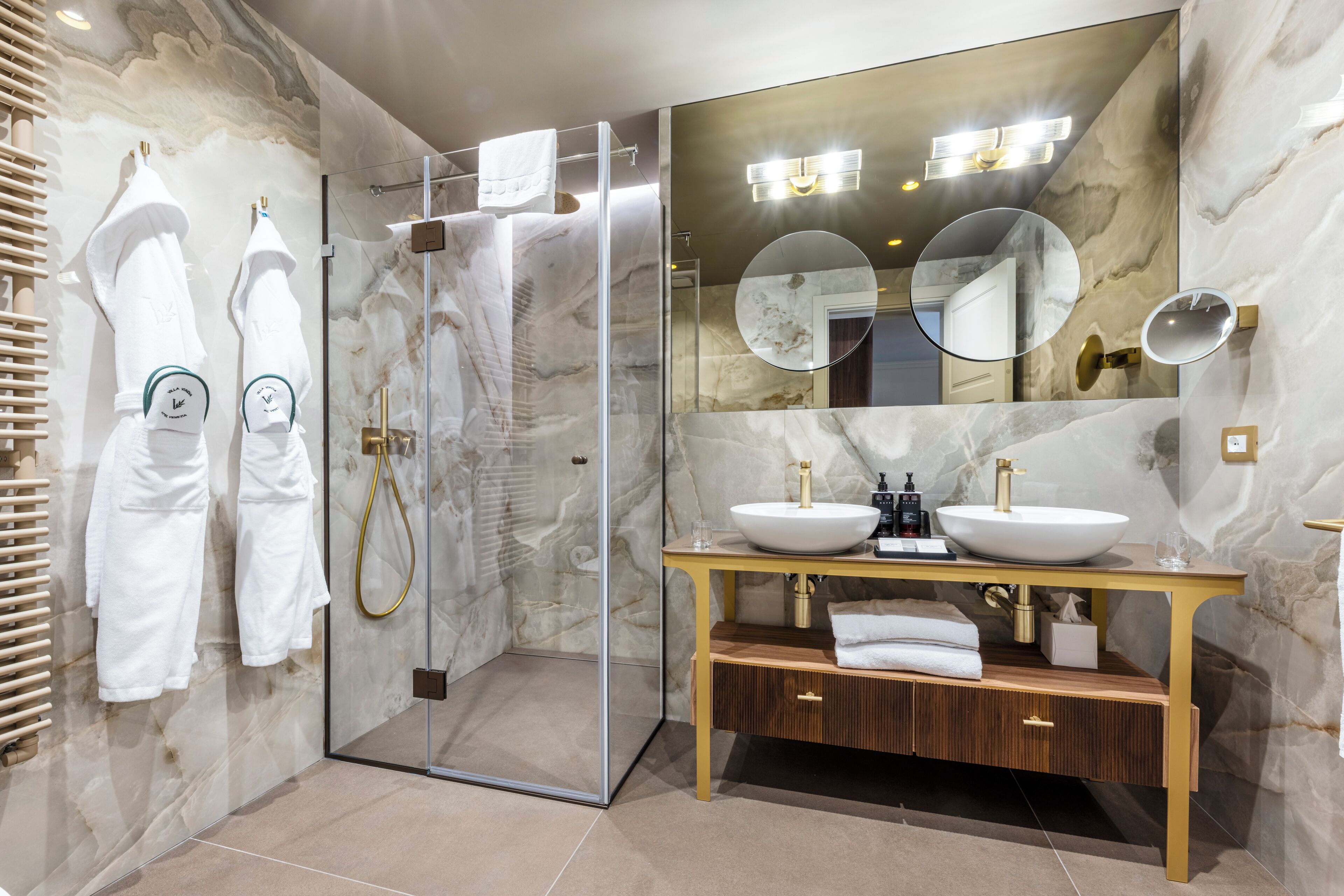 A modern bathroom with a glass-enclosed shower, two sinks with gold fixtures, and marble walls.