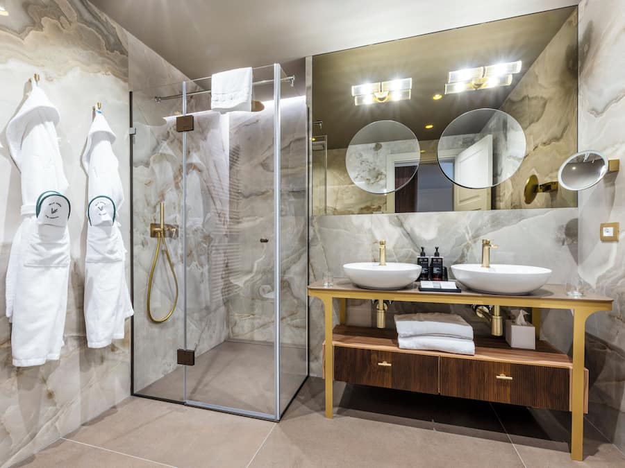 A modern bathroom with a glass-enclosed shower, two sinks with gold fixtures, and marble walls.