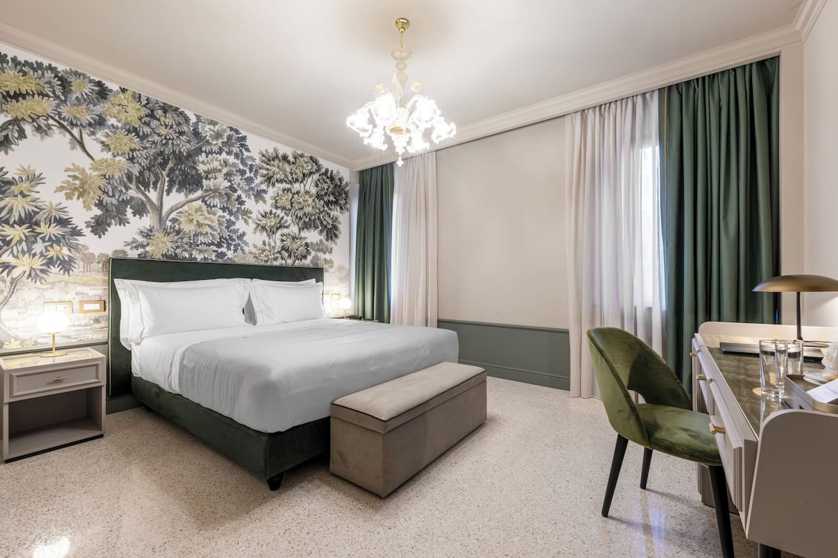 A hotel room with a large bed, a desk with a chair, a nightstand, a bench, and a wall with a floral mural.