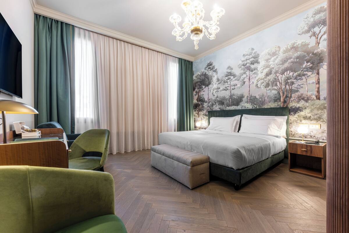A hotel room with a large bed, a desk with a lamp, a green armchair, and a painting of trees on the wall.