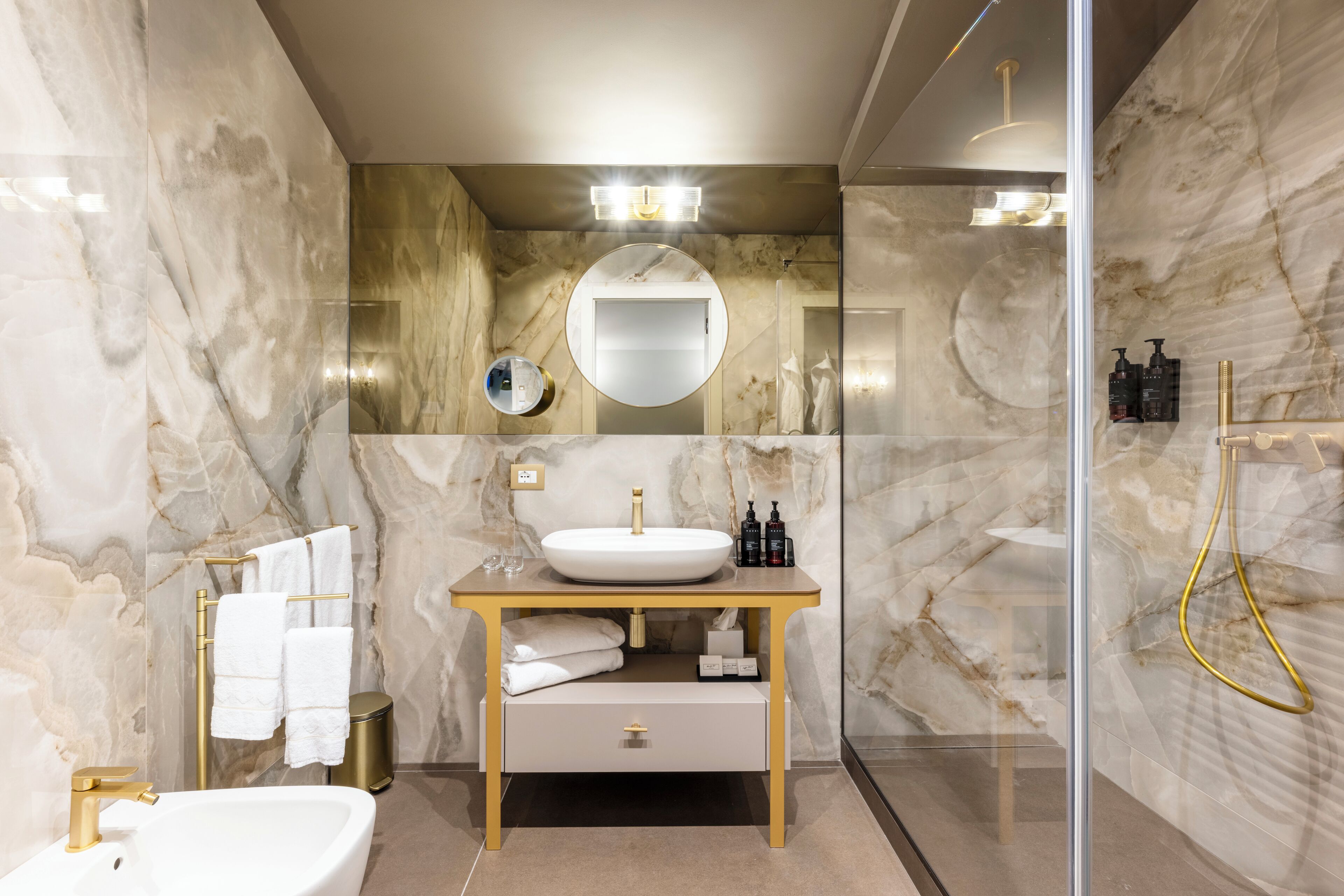Un baño moderno con pared de mármol, un espejo redondo, un lavamanos blanco y un grifo dorado.