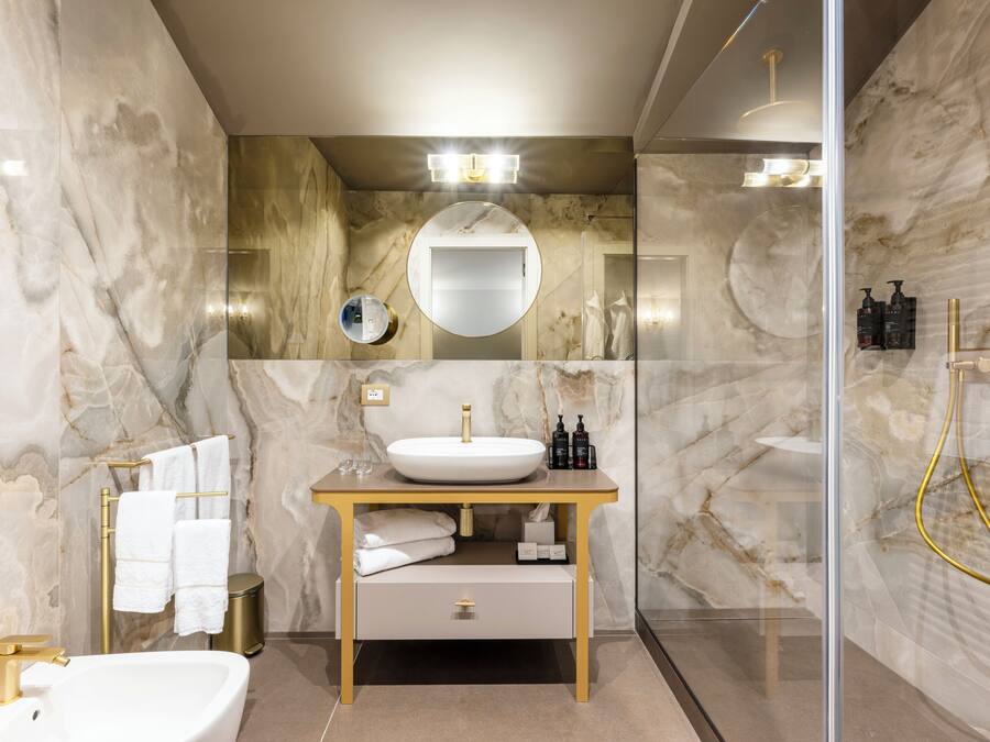 Un baño moderno con pared de mármol, un espejo redondo, un lavamanos blanco y un grifo dorado.