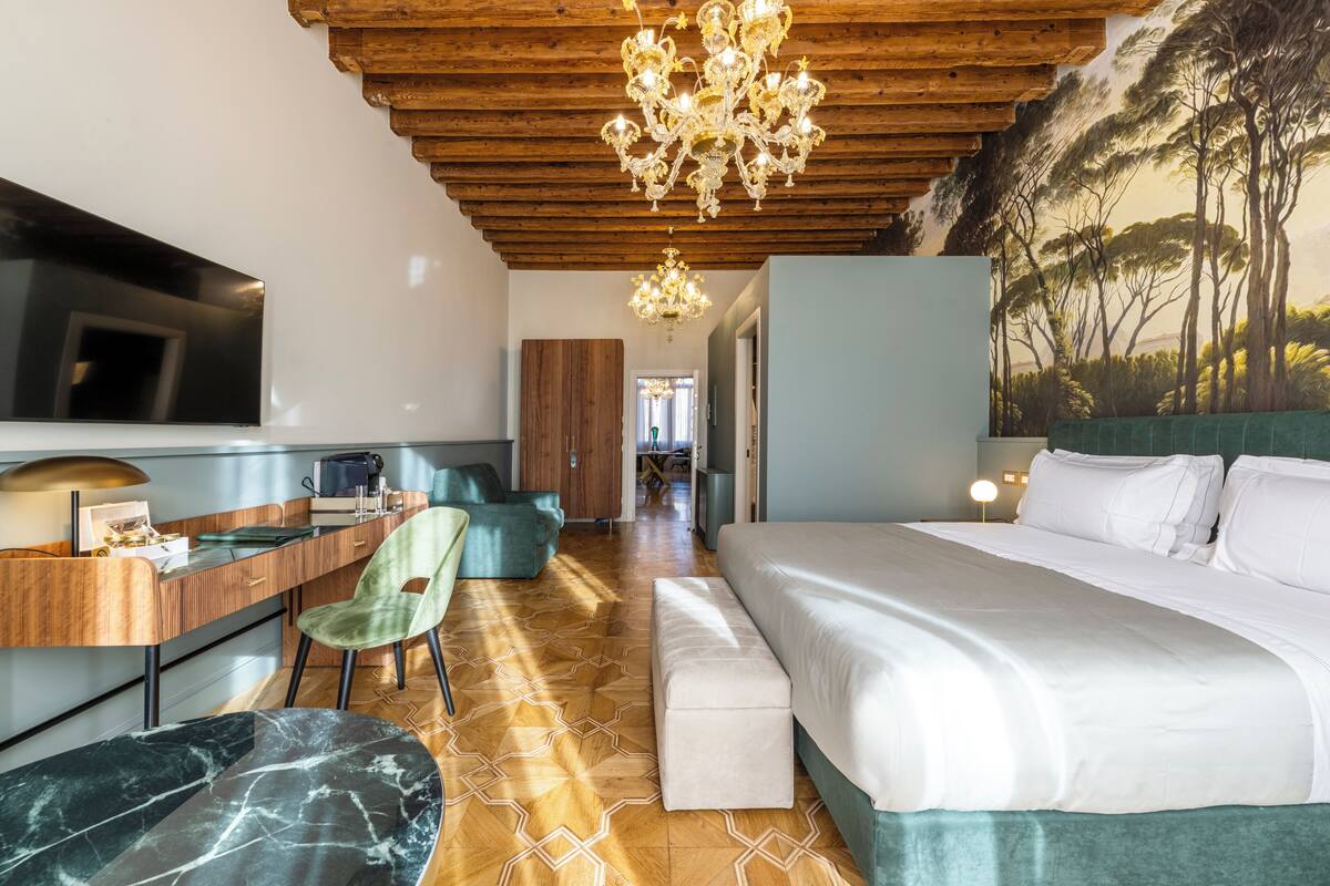 A modern hotel room with a wooden ceiling, a large bed, a desk with a chair, and a mural of trees on the wall.