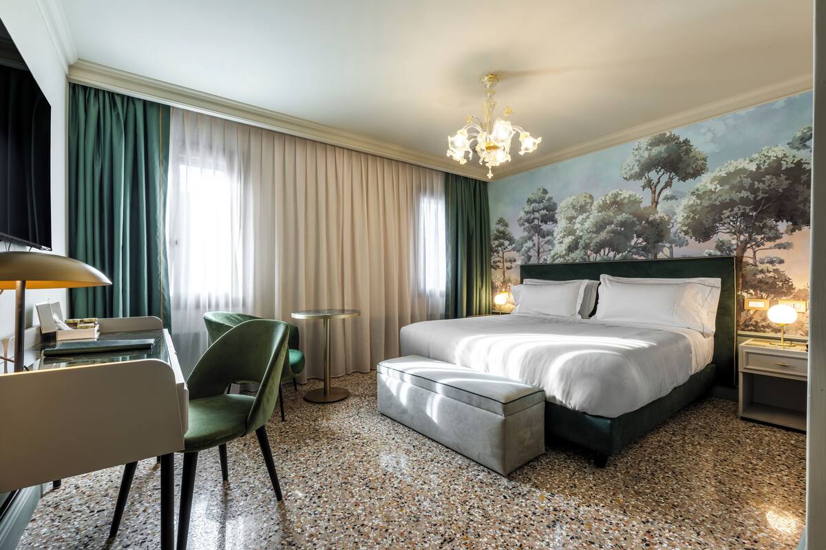 A hotel room with a bed, a desk, a chair, a small table, and a painting on the wall.