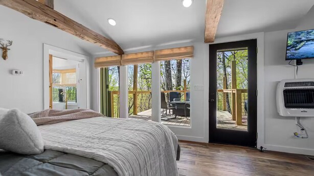 Un dormitorio con una cama grande, techo de madera, puerta corrediza de vidrio a una terraza y televisor montado.