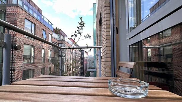 Uma mesa de madeira com cinzeiro transparente, com um edifício moderno ao fundo, com grandes janelas e céu claro.