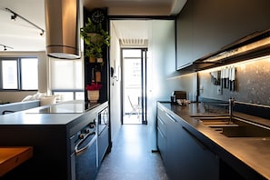 A modern kitchen with a central island, built-in oven, and a large window. - Gávea | 1 quarto, vista Cristo, prédio com infra - por Casapē Rio (Rio de Janeiro)