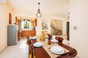 A dining area with a wooden table set for four, a kitchen in the background, and a staircase leading to another floor. - Villa Phedra - Three Bedroom Villa, Sleeps 6 (Kefalas)