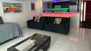 A modern living room with a sofa set, a coffee table, and a kitchen area with a neon-lit countertop.
