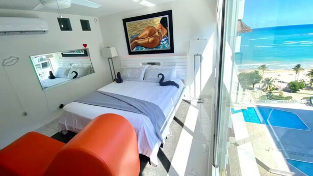 A hotel room with a bed, a TV, a window with a view of the beach and ocean, and a ceiling fan.