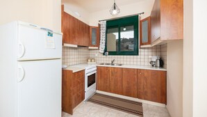 A compact kitchen with wooden cabinets, a white refrigerator, a small oven, and a window with a green frame. - Villa Yvonne - Three Bedroom Villa, Sleeps 6 (Kefalas)