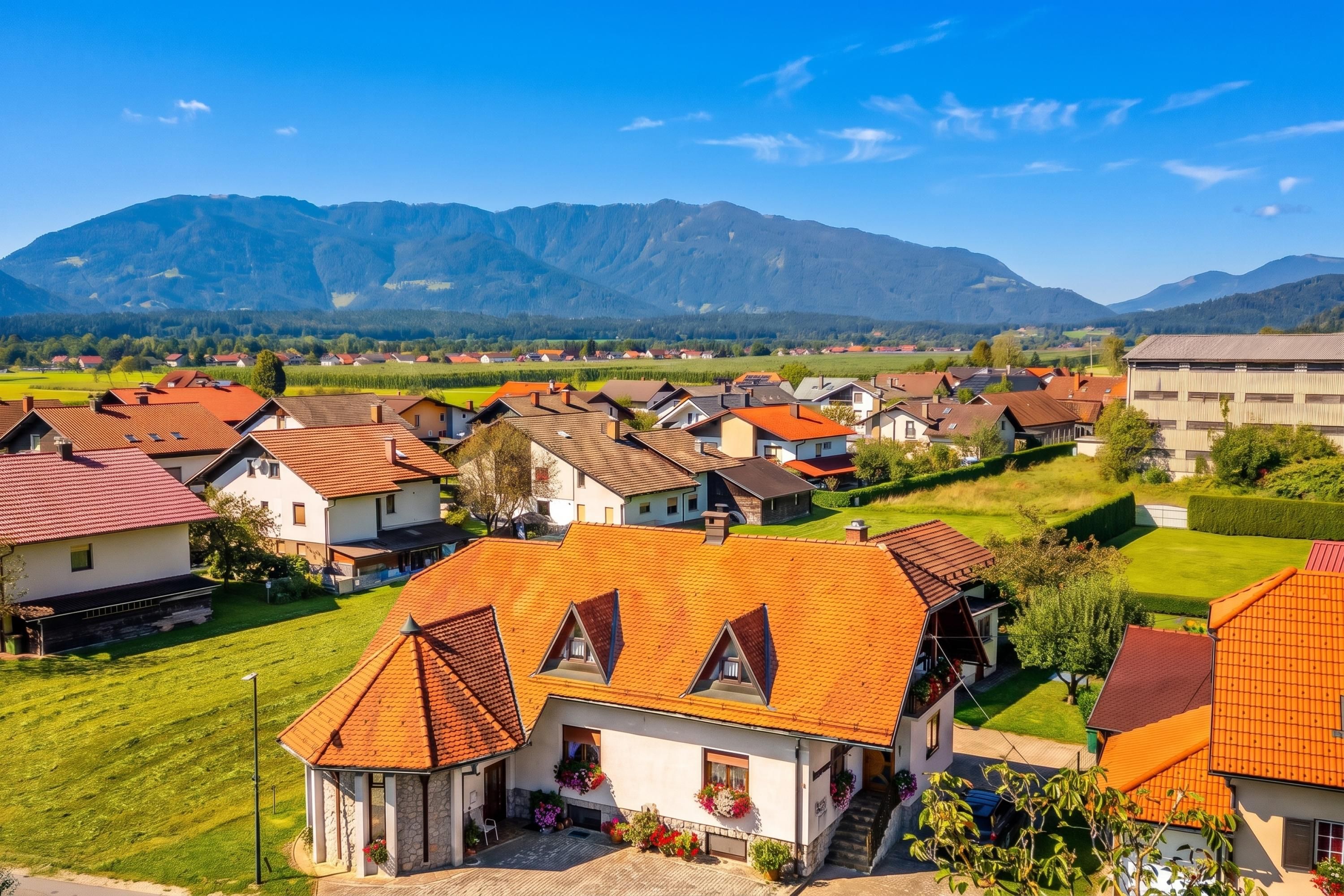 Ein Wohngebiet mit Häusern und roten Ziegeldächern, umgeben von grünen Feldern und Bergen im Hintergrund.