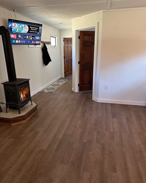 Una sala de estar con una estufa de leña, un televisor montado que muestra un servicio de streaming, y un pasillo que conduce a una puerta.