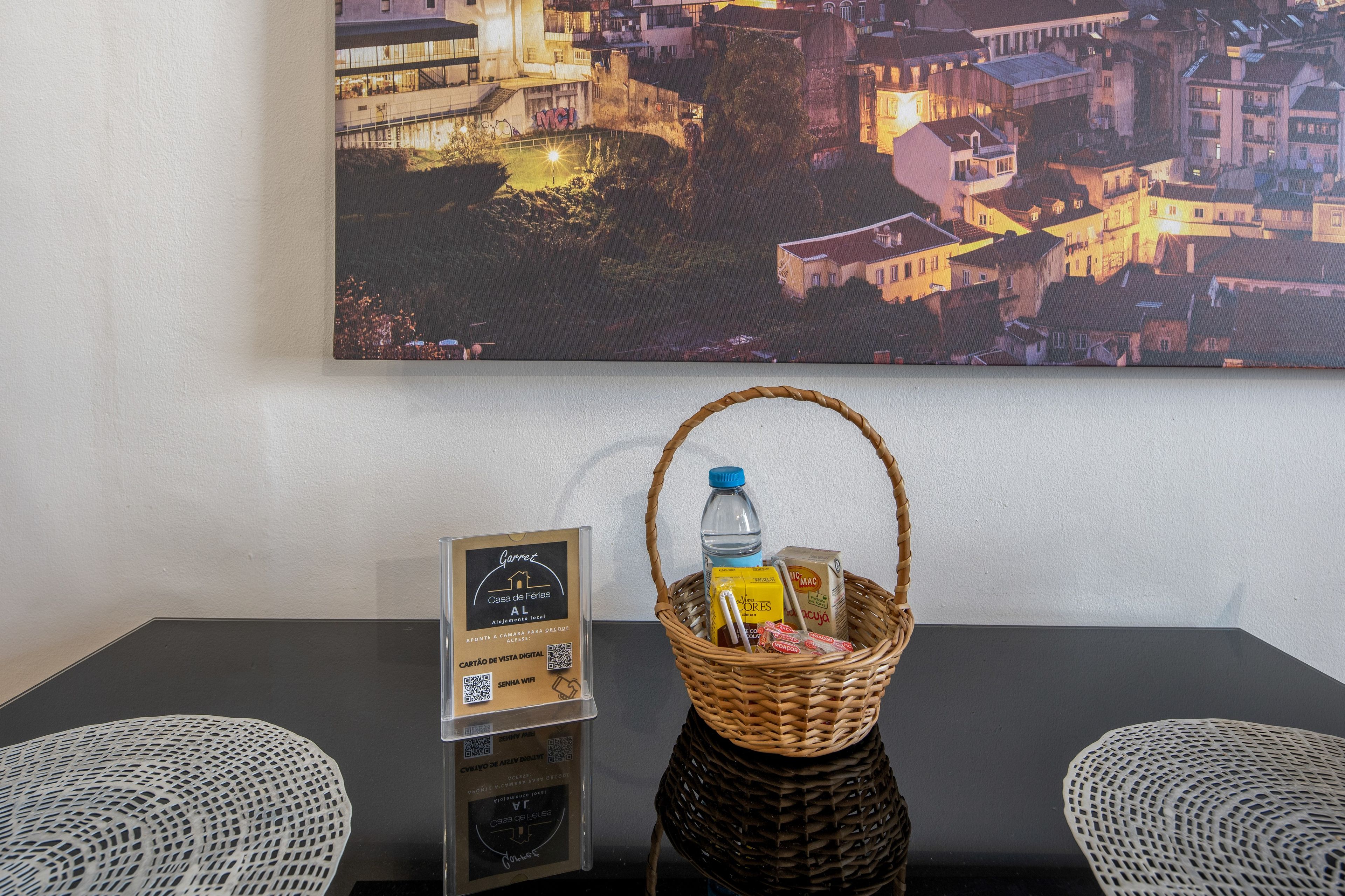 A hotel room with a table featuring a QR code sign, a wicker basket with a bottle and snacks, and a cityscape painting on the wall.
