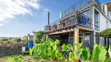 Een modern herenhuis met twee verdiepingen, een balkon, een stenen muur en een tuin met grote, weelderige planten.