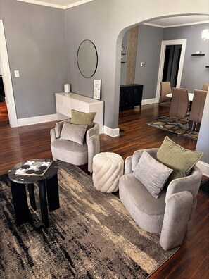 A modern living room with a grey wall, a round mirror, a white console table, and a dining area with brown chairs.