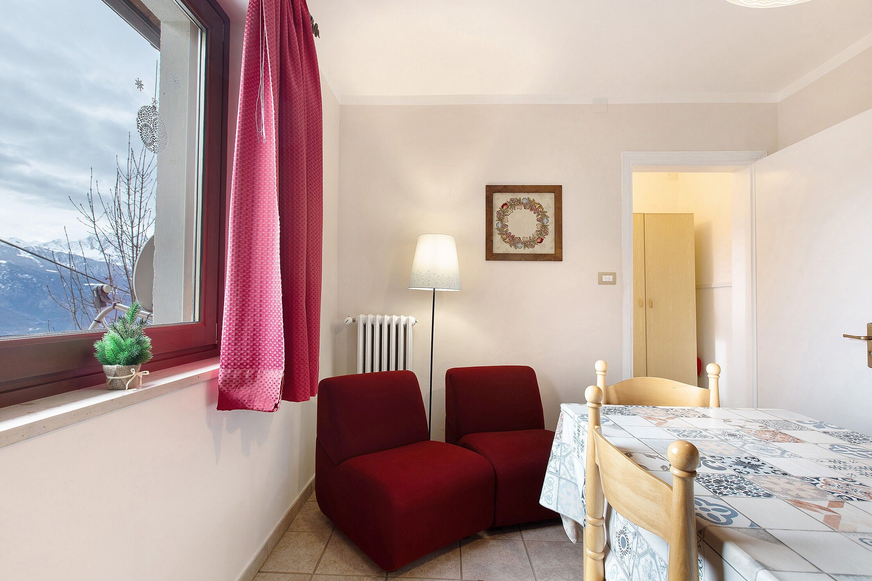 A small room with a window, a radiator, a table with a patterned tablecloth, two red chairs, and a small potted plant.
