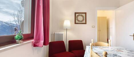 A small room with a window, a radiator, a table with a patterned tablecloth, two red chairs, and a small potted plant.