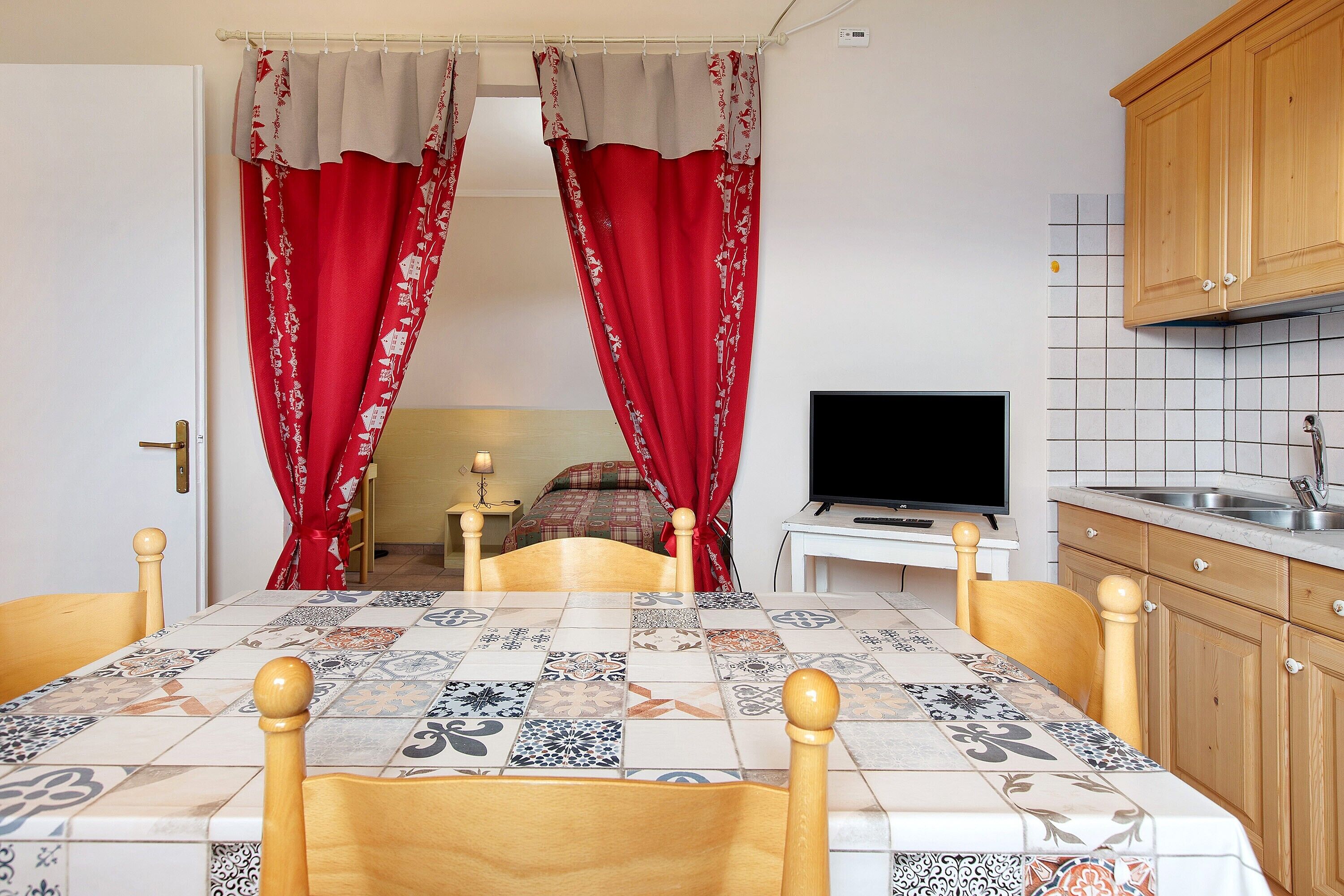 Una habitación con una mesa enlosada, sillas de madera, un televisor y una zona de cocina con armarios de madera y salpicadero enlosado.