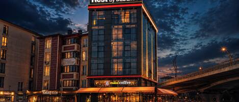 Un hotel con un llamativo letrero de 'Rest by DEDEMAN', una cafetería llamada 'mikel coffee' en la parte inferior, y una calle concurrida con estelas de luz.
