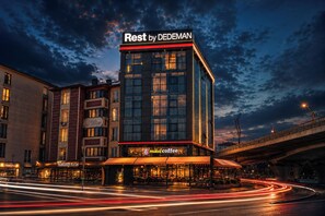 Un hotel con un cartello ben visibile 'Rest by DEDEMAN', una caffetteria chiamata 'mikel coffee' sottostante e una strada trafficata con scie luminose.