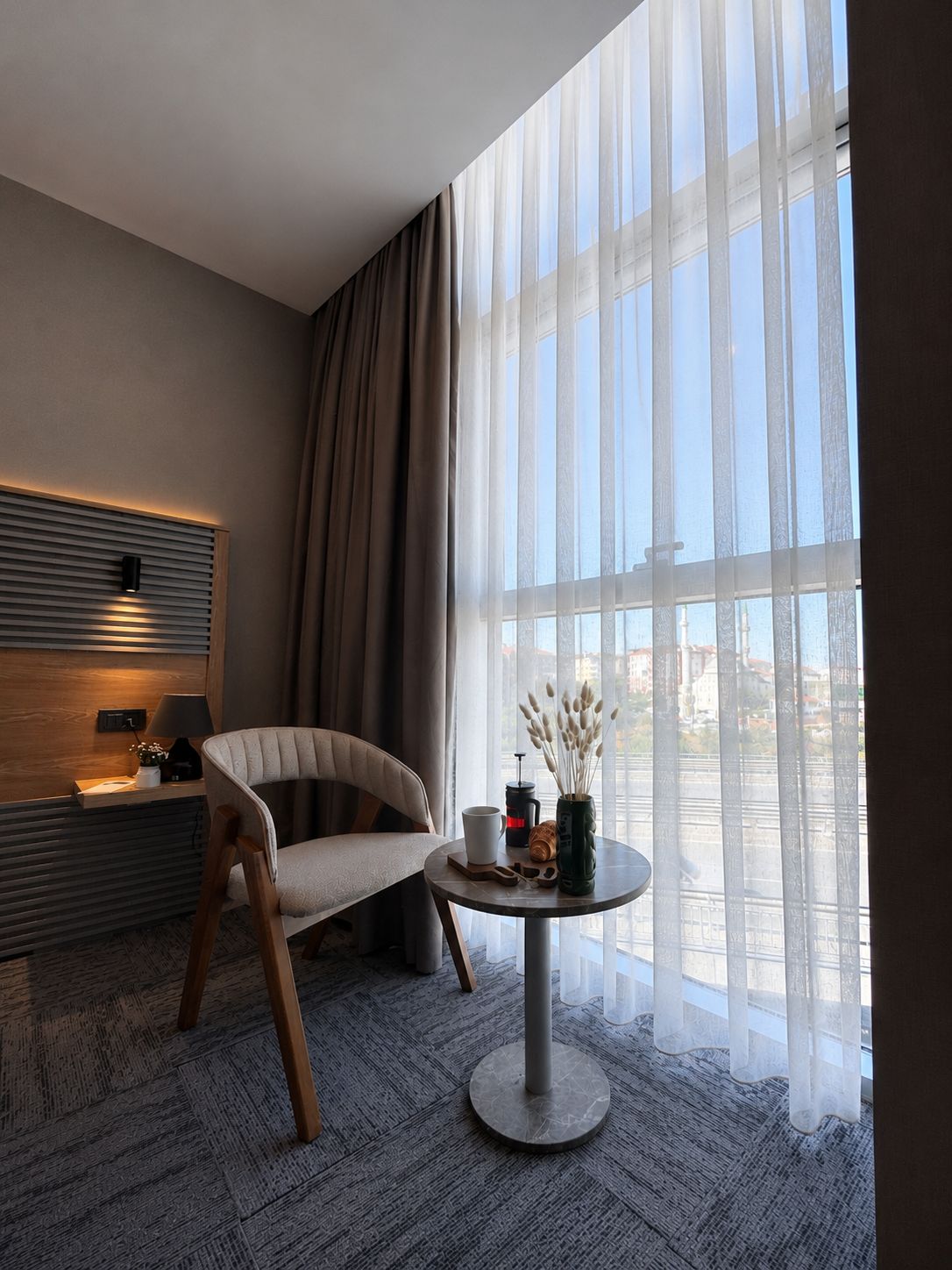 A modern hotel room with a large window, a round table with a vase of pampas grass, a chair, and a bedside table with a lamp.