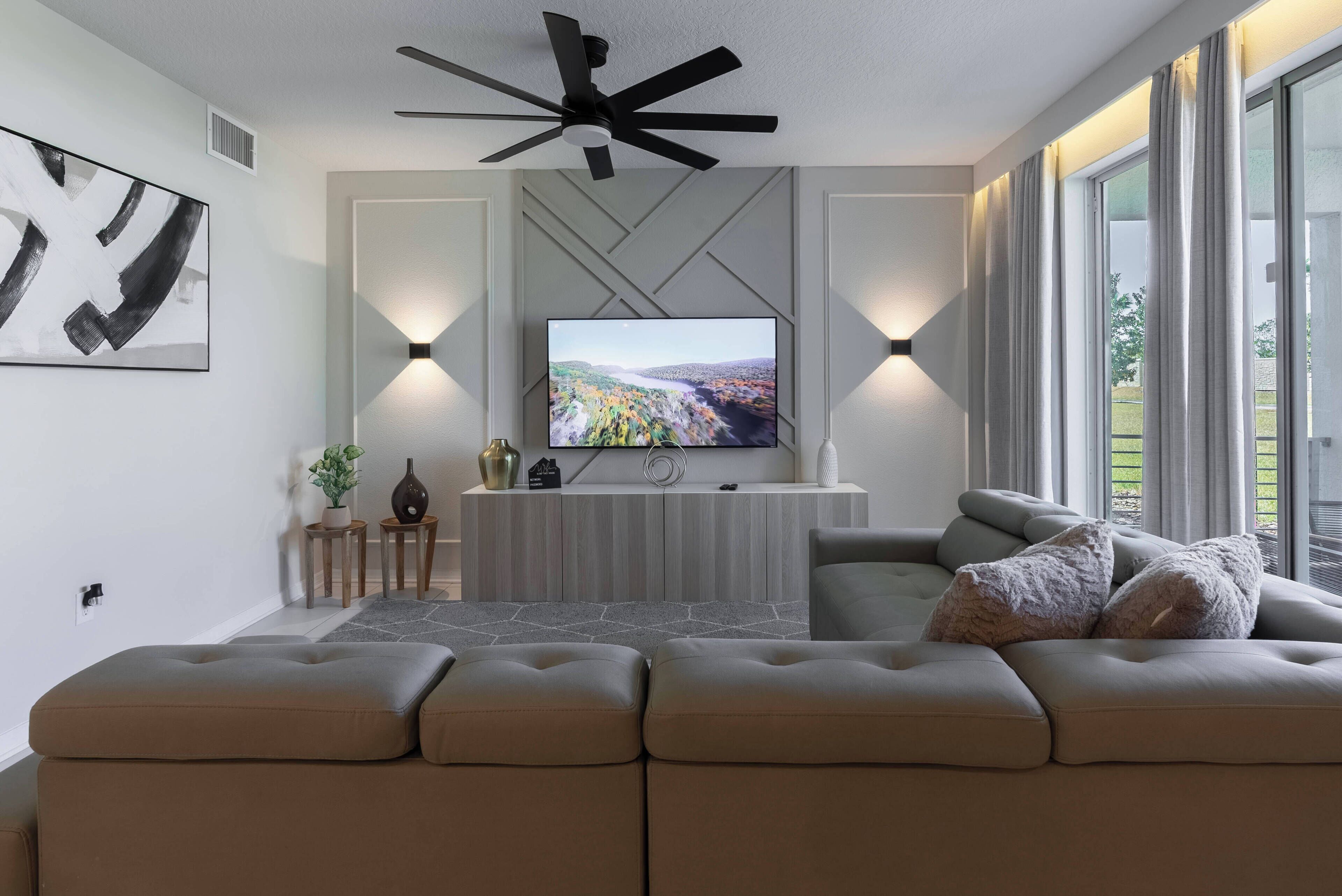 A modern living room with a large flat-screen TV, a ceiling fan, and a sofa with cushions.