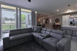 Uma sala de estar moderna com um sofá seccional cinza, uma área de jantar com mesa redonda e cadeiras, e uma cozinha com geladeira de aço inoxidável.
