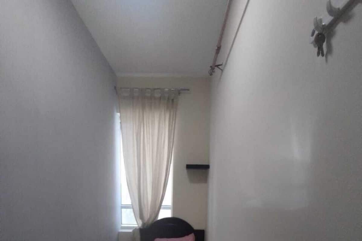 A narrow, dimly lit room with a single bed, a window with sheer curtains, and a small shelf.