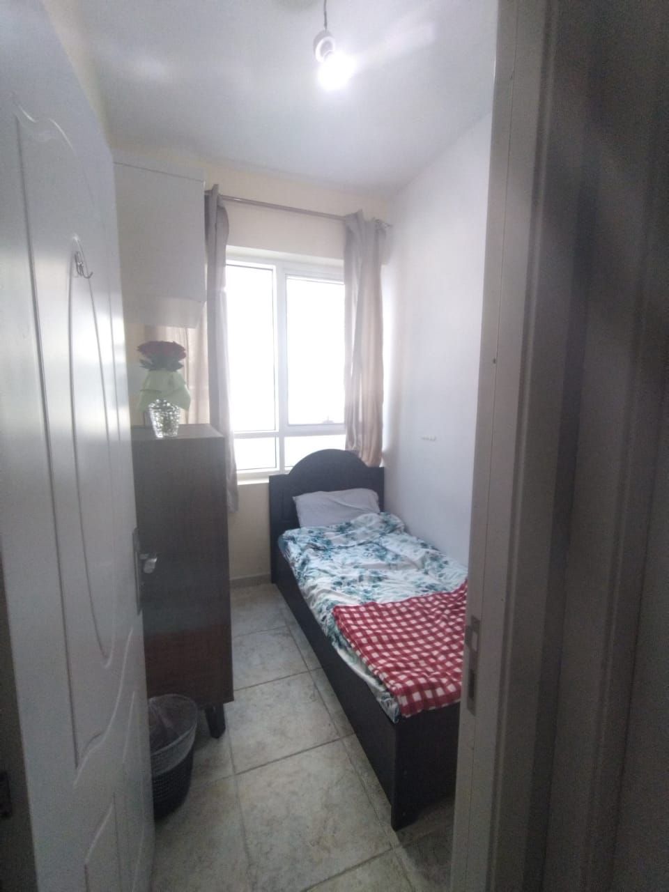Une petite pièce étroite, avec un lit simple, une commode et une fenêtre avec des rideaux.