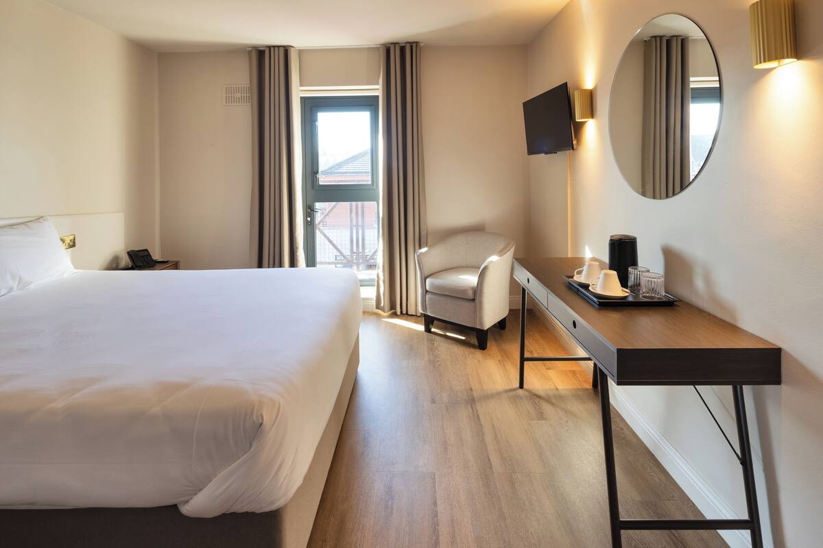 Habitación de hotel con una cama grande, un escritorio con silla, un televisor de pantalla plana, un espejo y una ventana con cortinas.