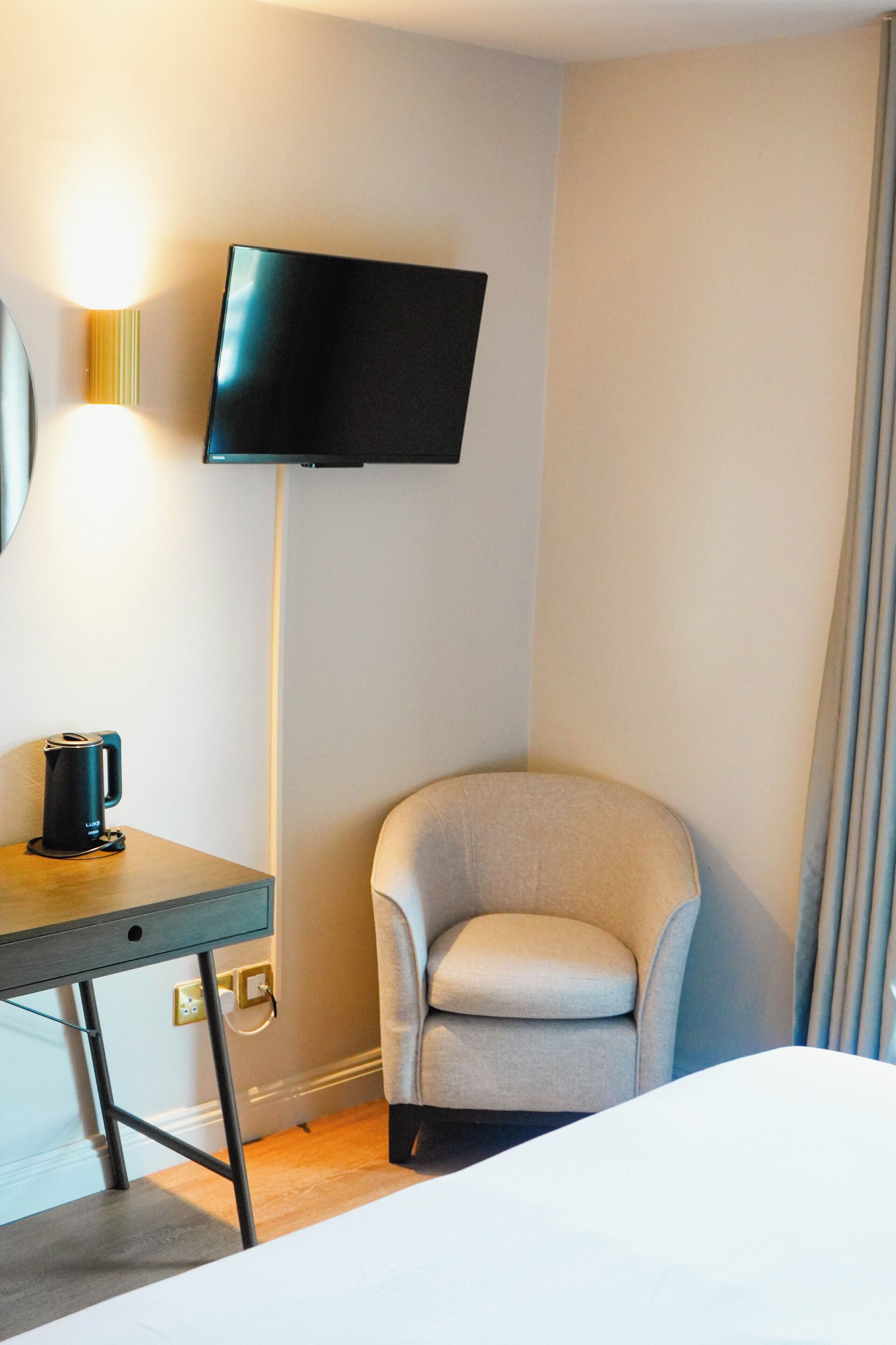 A hotel room with a bed, a desk with a kettle, a chair, a TV mounted on the wall, and a curtain.