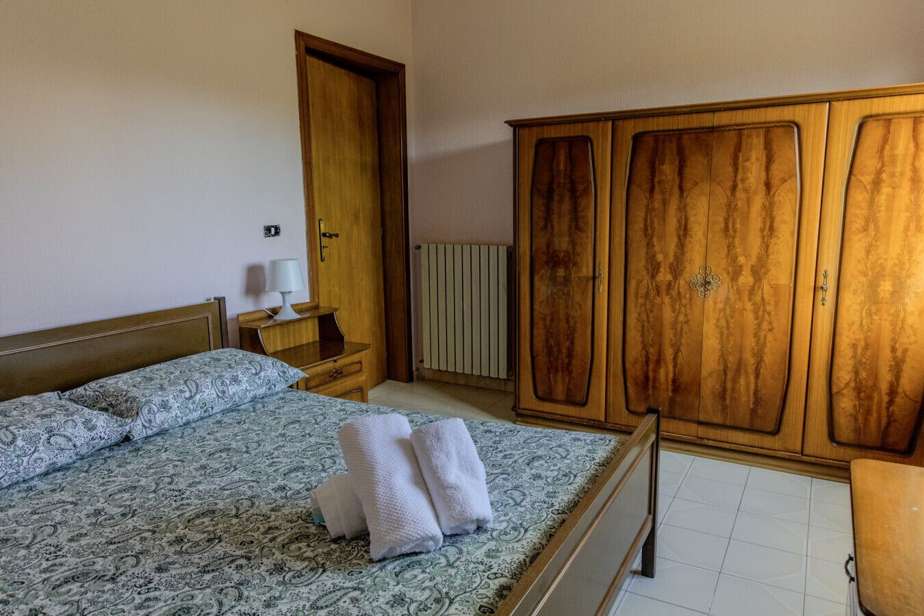 Ein Schlafzimmer mit einem Bett, einem Holzschrank, einem Nachttisch mit Lampe und einer Heizung.