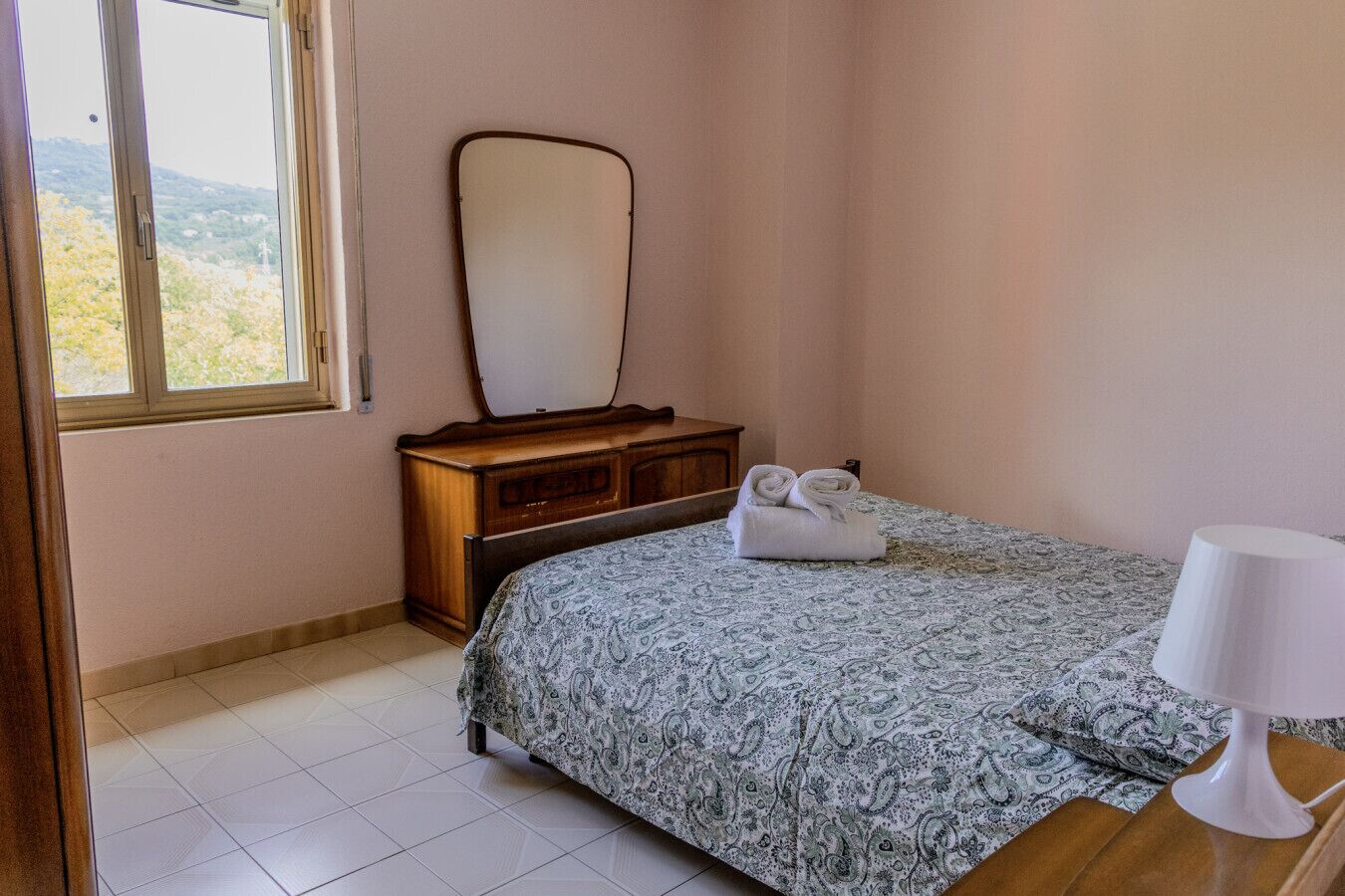 Ein Schlafzimmer mit einem Bett, einer Holzkommode, einem Spiegel und einem Fenster mit Blick auf Bäume.