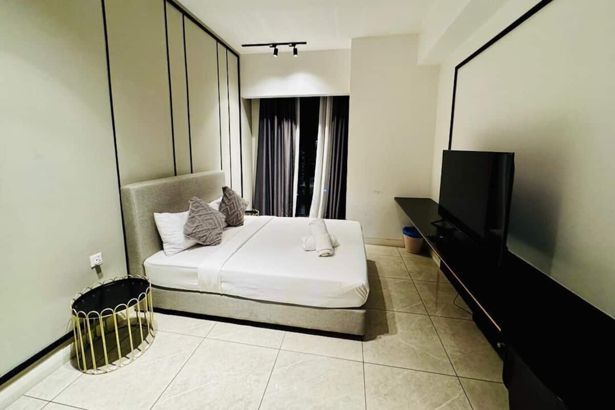 A modern hotel room with a bed, a television, a small trash bin, and a window with curtains.