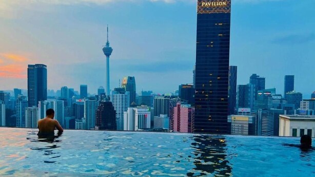 Um homem em uma piscina infinita com vista para uma paisagem urbana, com edifícios notáveis, incluindo um arranha-céu alto identificado como "Pavilion".