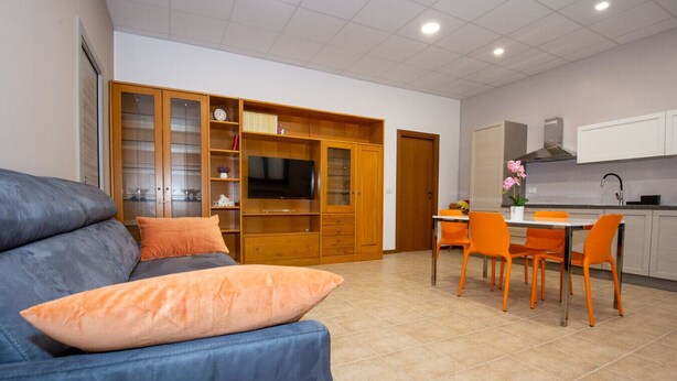A modern living room with a blue sofa, orange cushions, a wooden entertainment unit, and a dining area with orange chairs and a white table. - The Orchid - Francavilla al Mare (Francavilla al Mare)