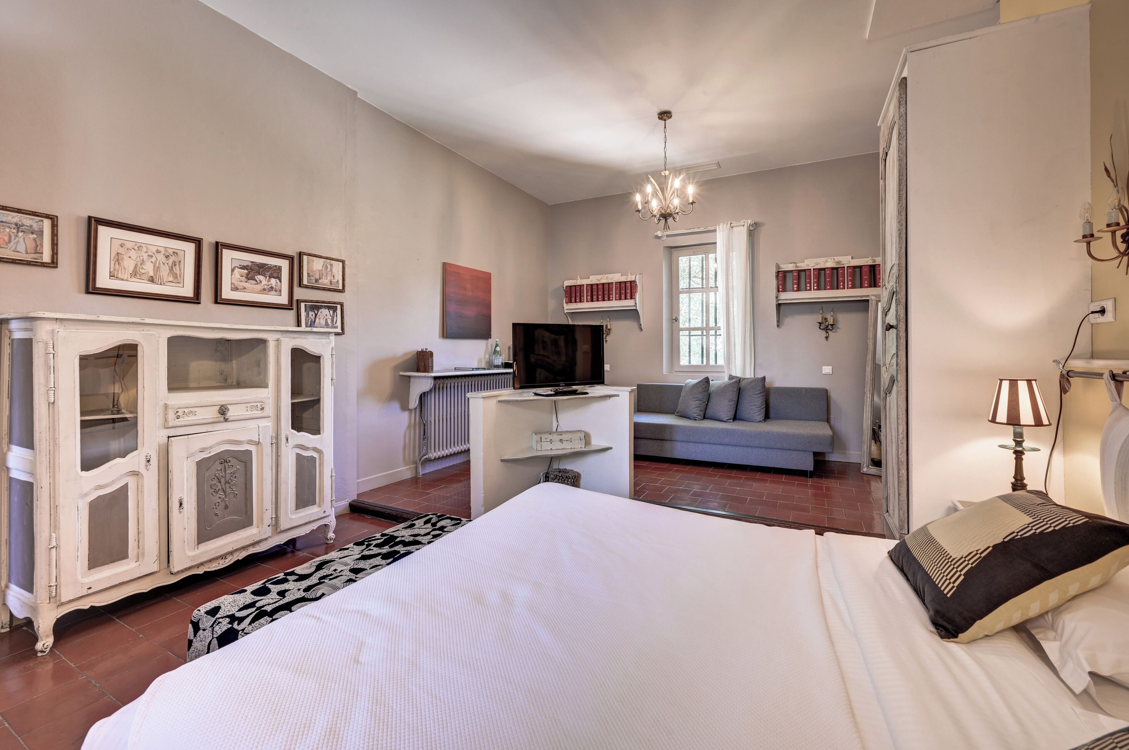 A bedroom with a large bed, a white wardrobe, a grey sofa, a television, and a bookshelf.
